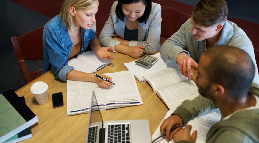 Group of four people collaborating at table reviewing documents and charts with laptops and coffee, discussing exam preparation strategy