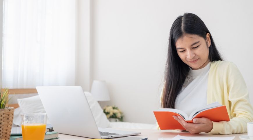Woman in yellow cardigan reading red book while working at laptop desk with glass of juice and plant nearby